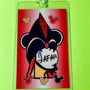 May include: A red and white luggage tag with a cartoon image of Jafar from Aladdin. The image is surrounded by Mickey Mouse ears. The text "JAFFAR" is written in white on a white circle.