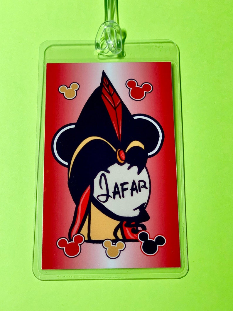 May include: A red and white luggage tag with a cartoon image of Jafar from Aladdin. The image is surrounded by Mickey Mouse ears. The text "JAFFAR" is written in white on a white circle.