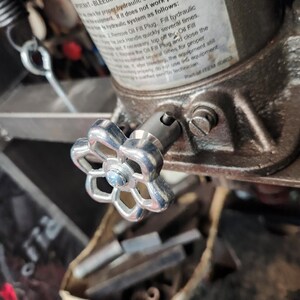 May include: Close-up of a silver-colored valve with a flower-shaped handle attached to a hydraulic jack. The jack has a label with instructions for bleeding the hydraulic system. The background is blurred, showing other mechanical parts.