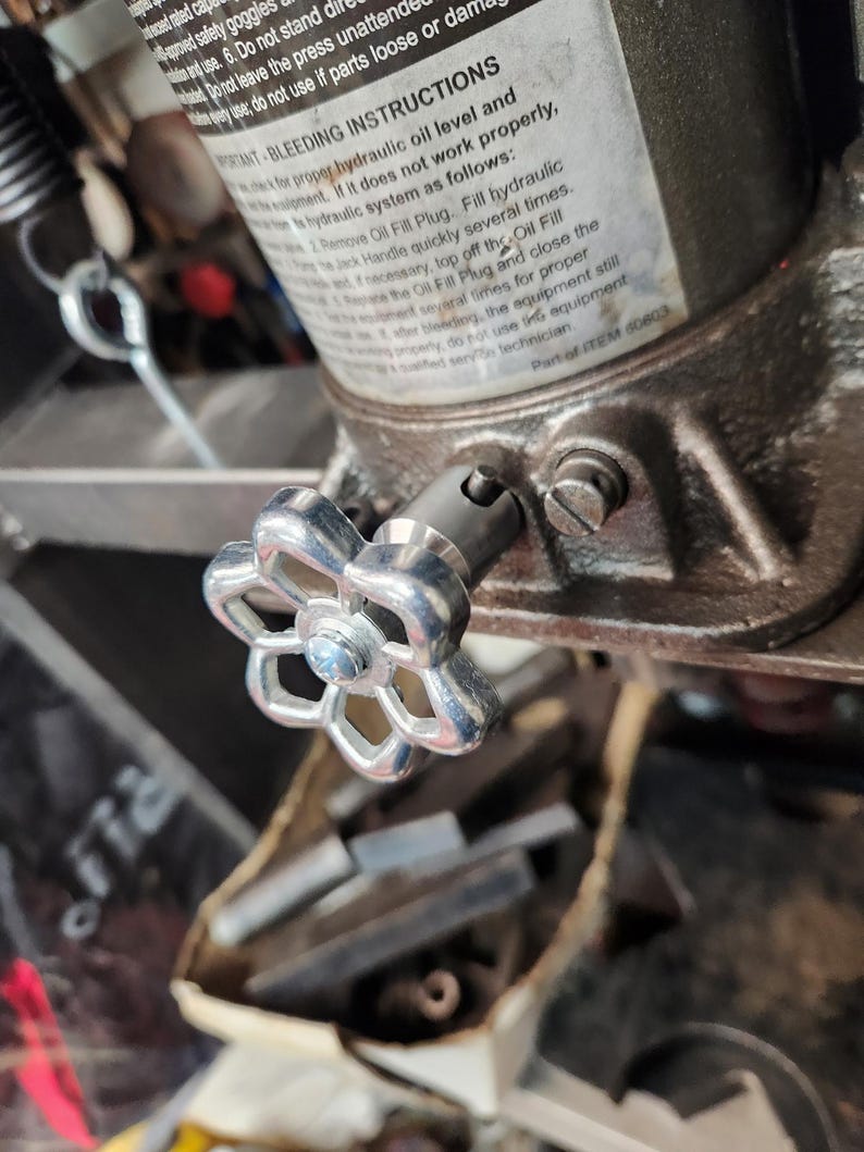 May include: Close-up of a silver-colored valve with a flower-shaped handle attached to a hydraulic jack. The jack has a label with instructions for bleeding the hydraulic system. The background is blurred, showing other mechanical parts.