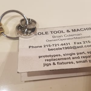 May include: A brass-colored bolt keychain with a silver key ring and two silver washers. A business card with the text "COLE TOOL & MACHINE" is attached to the key ring. The card lists contact information and services.