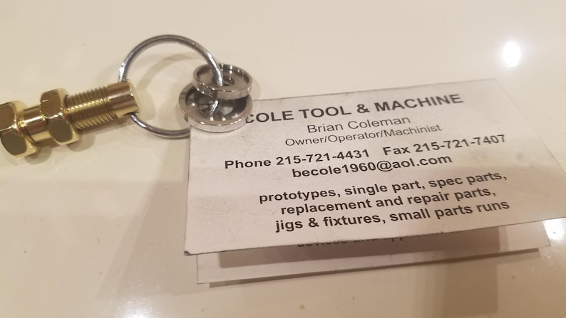 May include: A brass-colored bolt keychain with a silver key ring and two silver washers. A business card with the text "COLE TOOL & MACHINE" is attached to the key ring. The card lists contact information and services.