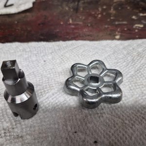 May include: Two metal components, a valve stem and a flower-shaped handle, rest on a white paper towel. The valve stem is cylindrical with a square top. The handle has a six-petal design with a square center opening. Both are silver in color.