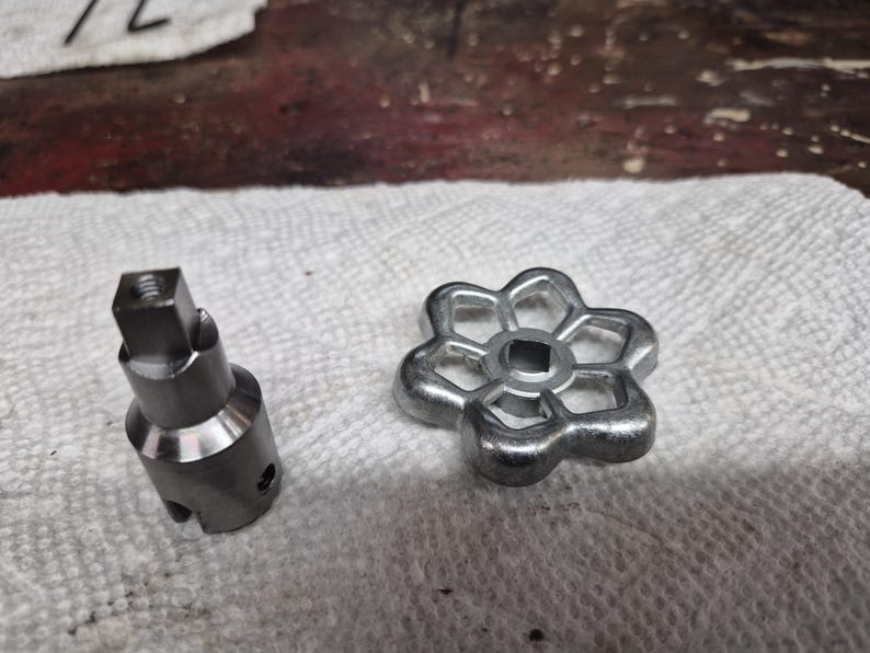May include: Two metal components, a valve stem and a flower-shaped handle, rest on a white paper towel. The valve stem is cylindrical with a square top. The handle has a six-petal design with a square center opening. Both are silver in color.