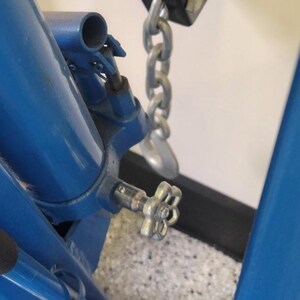 May include: Close-up of a blue metal mechanical device with a silver chain and a flower-shaped handle. The device is bolted together and appears to be part of a larger machine. The background is a speckled gray and white surface.