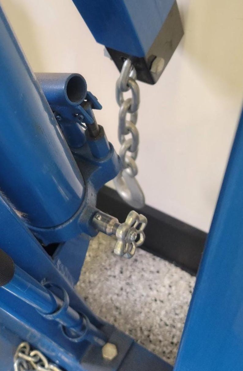 May include: Close-up of a blue metal mechanical device with a silver chain and a flower-shaped handle. The device is bolted together and appears to be part of a larger machine. The background is a speckled gray and white surface.