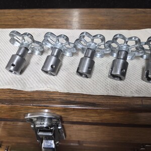 May include: Five silver-colored valve handles with a flower-like design on top, arranged in a row on a white paper towel. The handles have a cylindrical body and are made of metal. The background is a wooden surface.