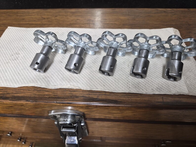 May include: Five silver-colored valve handles with a flower-like design on top, arranged in a row on a white paper towel. The handles have a cylindrical body and are made of metal. The background is a wooden surface.
