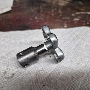 May include: A metallic silver-colored mechanical component with a butterfly-shaped handle. The component has a cylindrical body with a threaded hole and a shaft. It rests on a white, textured surface, suggesting a workshop or industrial setting.