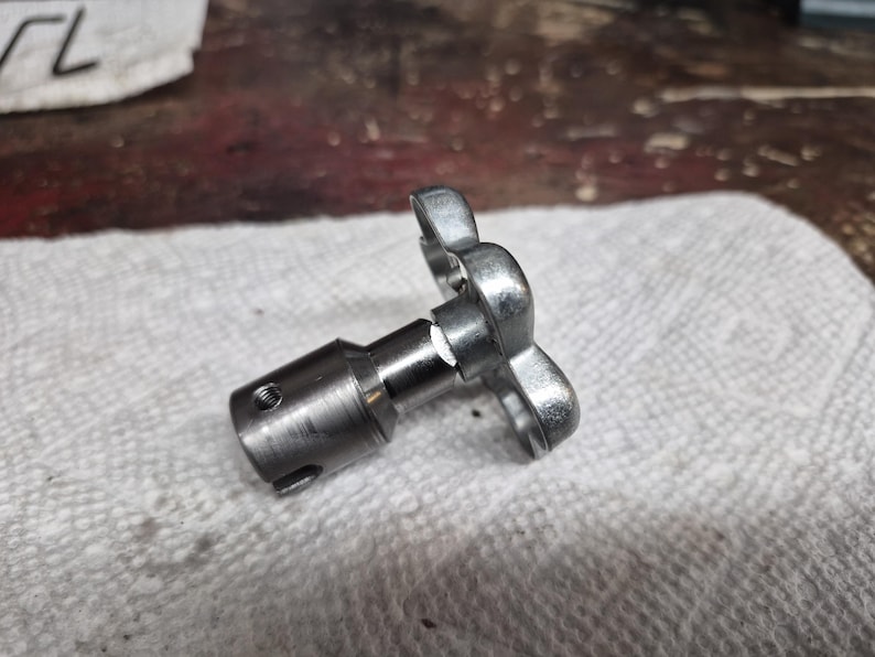 May include: A metallic silver-colored mechanical component with a butterfly-shaped handle. The component has a cylindrical body with a threaded hole and a shaft. It rests on a white, textured surface, suggesting a workshop or industrial setting.