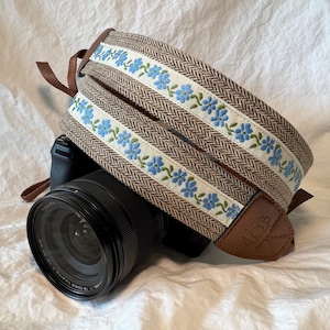 May include: A brown and white camera strap with blue floral accents. The strap is made of tweed fabric and has a brown leather patch with the word "TOB" on it.