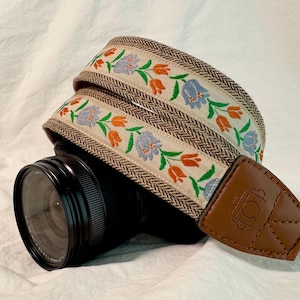 May include: A brown and white camera strap with a floral pattern. The strap has a brown leather end with a camera logo.