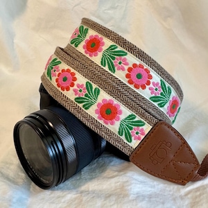 May include: A camera strap with a floral embroidered design. The strap features a cream-colored band with pink and red flowers and green leaves. The edges are brown with a herringbone pattern. A brown leather end piece is attached to the camera.