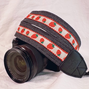 May include: A black camera strap with a white and red strawberry pattern. The strap is made of a woven fabric and has a leather camera attachment.