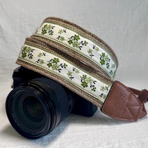 May include: A camera strap with a floral design. The strap features a white fabric with green floral embroidery and a brown herringbone border. The strap is attached to a camera with a brown leather end piece.