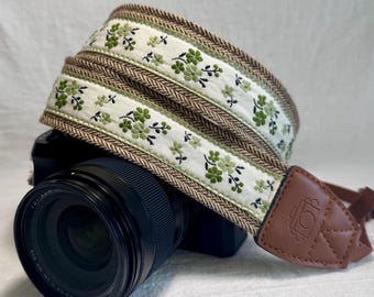 May include: A camera strap with a floral design. The strap features a white fabric with green floral embroidery and a brown herringbone border. The strap is attached to a camera with a brown leather end piece.