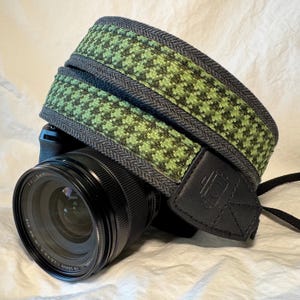 May include: A green and gray camera strap with a black leather strap and a camera lens attached.