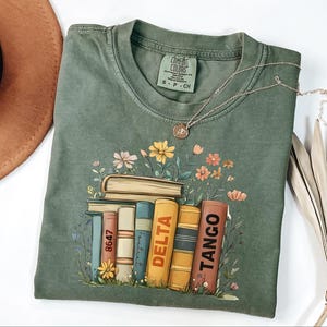 May include: Olive green Comfort Colors t-shirt with a floral and book design. The graphic features a stack of books with the words "Delta" and "Tango" on the spines. A silver necklace with a pendant is also visible.