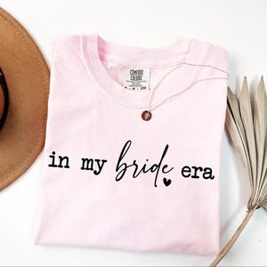 May include: A light pink t-shirt with the phrase "in my bride era" in black script. A delicate gold necklace with a pendant rests on the shirt. A brown hat and dried plant are in the background.
