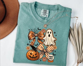 May include: A sage green t-shirt featuring a graphic print of a ghost, a jack-o'-lantern, a black crow, two large bows, flowers, stars, and a spiderweb. The text "TRICK OR TREAT" is printed on a small crystal ball graphic on the shirt.