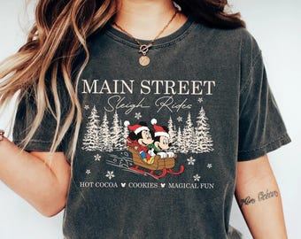 May include: Dark gray t-shirt featuring a graphic of Mickey and Minnie Mouse in a sleigh, with the text "MAIN STREET Sleigh Rides". The design includes snowflakes and pine trees, with the words "HOT COCOA COOKIES MAGICAL FUN" below.