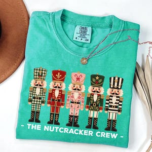 May include: A mint green short-sleeve t-shirt featuring a graphic of five nutcracker figures in various colors and patterns. Below the figures, the text reads THE NUTCRACKER CREW. The shirt has a Comfort Colors brand label at the inner neckline.