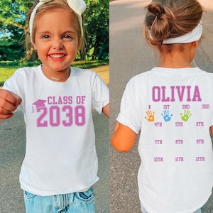 May include: White t-shirt with the text "CLASS OF 2038" in pink. The back of the shirt has the name "OLIVIA" and handprints indicating grade levels from K to 12th grade. The shirt is worn by a young child.