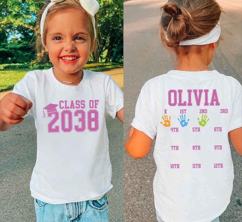 May include: White t-shirt with the text "CLASS OF 2038" in pink. The back of the shirt has the name "OLIVIA" and handprints indicating grade levels from K to 12th grade. The shirt is worn by a young child.