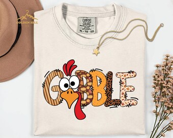 May include: A cream-colored short-sleeve t-shirt featuring a graphic of a turkey face integrated into the word GOBBLE. The letters have various patterns including stripes, polka dots, leopard print, and autumn leaves. The shirt has a Comfort Colors label.