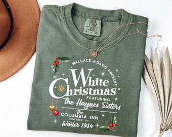 May include: Olive green t-shirt with a graphic promoting "White Christmas" featuring The Haynes Sisters. The design includes holly, ornaments, and text: "Wallace & Davis Present", "The Columbia Inn, Pinetree, VT, Winter 1954". A gold necklace is draped across the shirt.