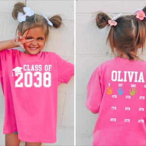 May include: A bright pink t-shirt with the text "CLASS OF 2038" in white. The back of the shirt has the name "OLIVIA" and handprints indicating school grades. A fun and personalized graduation shirt.
