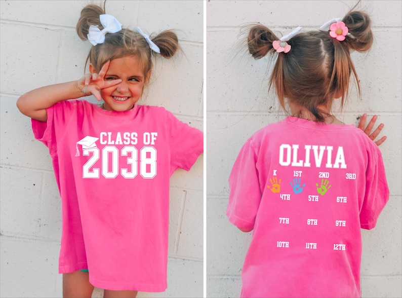 May include: A bright pink t-shirt with the text "CLASS OF 2038" in white. The back of the shirt has the name "OLIVIA" and handprints indicating school grades. A fun and personalized graduation shirt.