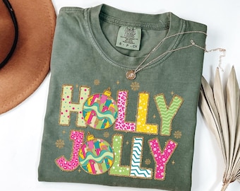 May include: Olive green t-shirt with the words "HOLLY JOLLY" in colorful, patterned letters and Christmas ornament graphics. A silver necklace with a pendant rests on the shirt. The shirt is folded.