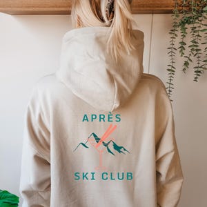 Könnte beinhalten: Beigefarbener Kapuzenpullover mit der Aufschrift "APRÈS SKI CLUB" in Türkis. Das Design zeigt ein Cocktailglas mit zwei rosa Strohhalmen und türkisfarbenen Bergen. Der Hoodie ist aus weichem Material.