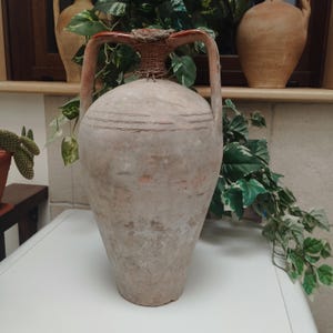 May include: A large, antique, beige-colored ceramic amphora with two handles and a woven neck. The vessel has horizontal etched lines and a weathered appearance, suggesting age and use. The handles are reddish-brown.