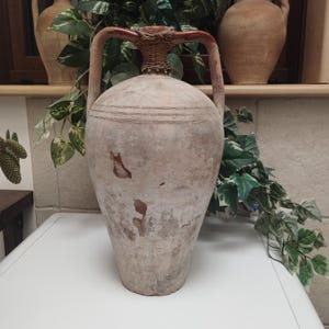 May include: A large, weathered, beige ceramic amphora with two handles and a woven neck. The vessel has a bulbous body and a narrow base. The amphora is decorated with horizontal lines and brown markings, suggesting age and use. It is a decorative item.