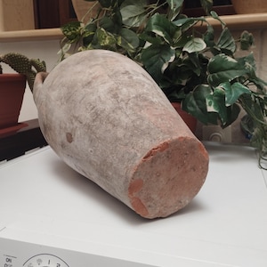 May include: A large, antique terracotta amphora with a weathered, textured surface. The vessel is a muted reddish-brown color with a lighter, dusty coating. The bottom is flat, and the top has a small handle. The amphora is likely used for storage.
