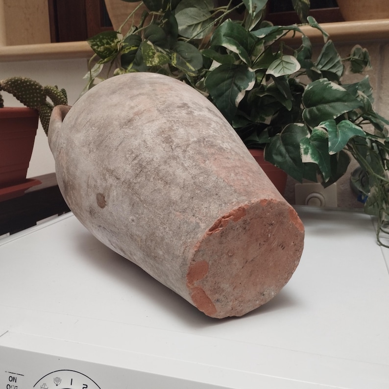 May include: A large, antique terracotta amphora with a weathered, textured surface. The vessel is a muted reddish-brown color with a lighter, dusty coating. The bottom is flat, and the top has a small handle. The amphora is likely used for storage.