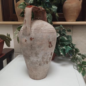 May include: A large, antique, beige-colored ceramic jug with a handle and a woven top. The jug has a wide base and tapers upwards, with horizontal lines near the top. The jug is likely used for decorative purposes.