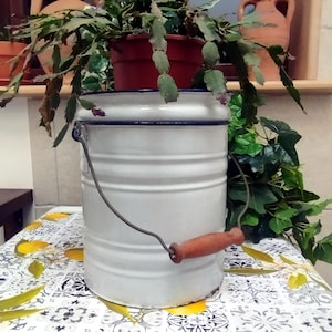 Puede incluir: Cubo de esmalte blanco con borde azul y asa de madera. El cubo está sobre una mesa con un patrón floral.