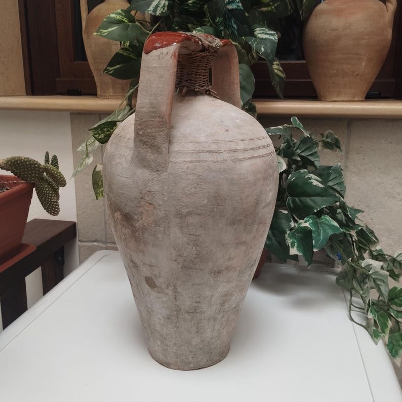 May include: A large, antique, beige-colored ceramic jug with two handles and a woven top. The jug has a rounded body and a narrow neck. The rim is painted red. The jug is likely used for storing liquids and has a rustic, aged appearance.