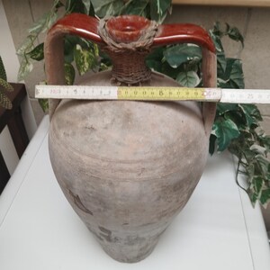 May include: A large, antique terracotta amphora with two handles and a decorative top. The amphora is approximately 20 inches wide. The handles are a reddish-brown color, and the top is woven. The amphora is a light brown color.