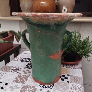 May include: A large, green ceramic vase with two handles and a wide, flat rim. The vase has a distressed, aged look with cracks and exposed reddish-brown clay. The rim and base are also reddish-brown. The vase is likely for decorative use, possibly for flowers.