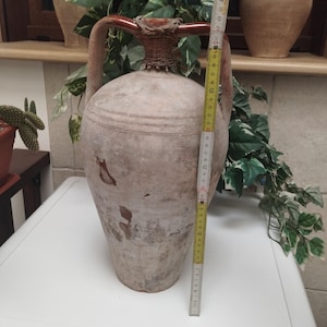 May include: A large, antique-style terracotta amphora with two handles and a woven neck. The amphora is approximately 30 inches tall. The vessel has a weathered, aged appearance with brown and beige tones, suggesting it is an old decorative piece.