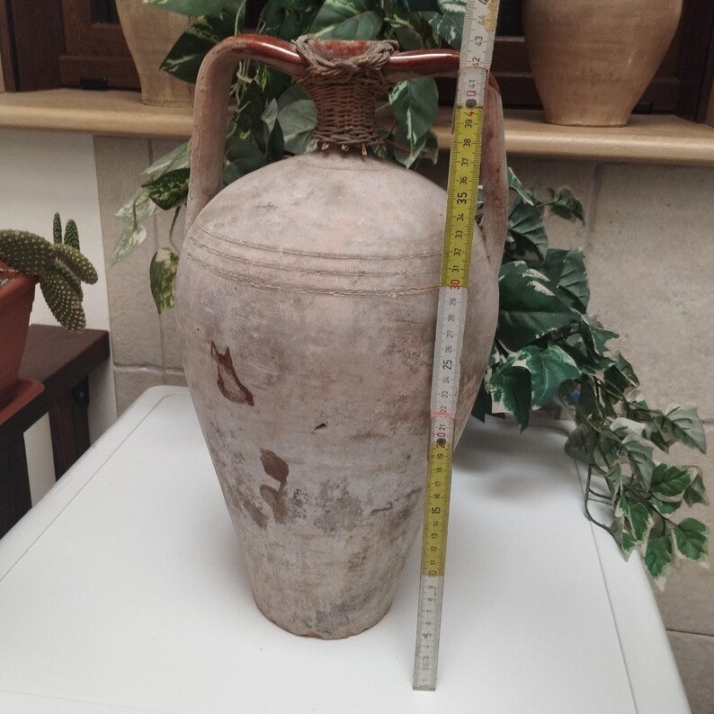 May include: A large, antique-style terracotta amphora with two handles and a woven neck. The amphora is approximately 30 inches tall. The vessel has a weathered, aged appearance with brown and beige tones, suggesting it is an old decorative piece.