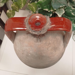 May include: A rustic, earthenware jug with a red, arched handle and a circular opening. The jug has a bulbous body and a weathered, textured surface. The handle is attached to the jug with a woven material.
