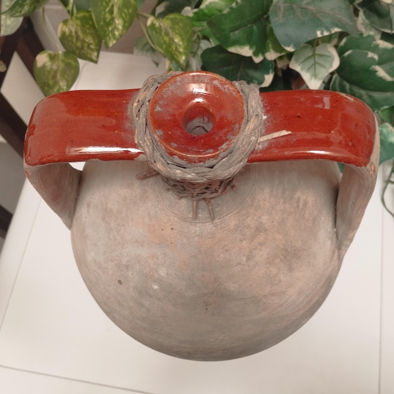May include: A rustic, earthenware jug with a red, arched handle and a circular opening. The jug has a bulbous body and a weathered, textured surface. The handle is attached to the jug with a woven material.