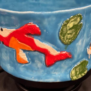 May include: A ceramic bowl with a light blue exterior, featuring raised relief koi fish and lily pads. The koi are painted in red, orange, and white, while the lily pads are green. The bowl is a decorative item.
