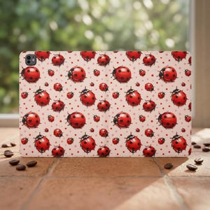 Pode incluir: Uma capa retangular para tablet com um padrão repetitivo de joaninhas vermelhas com pintas pretas sobre um fundo rosa claro. O design inclui pontos vermelhos menores intercalados entre as joaninhas, com um recorte para a câmara no canto.