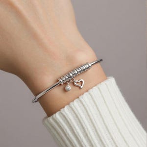 Könnte beinhalten: Ein silbernes Armreif-Armband mit gravierten Perlen. Ein Herz-Anhänger und ein kleiner, runder Anhänger mit einem zentralen Stein hängen daran. Das Armband wird am Handgelenk getragen, vor einem weißen Pullover.